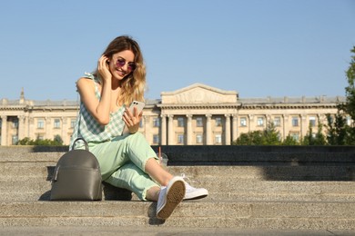 Photo of Young woman with stylish backpack and smartphone on stairs outdoors. Space for text