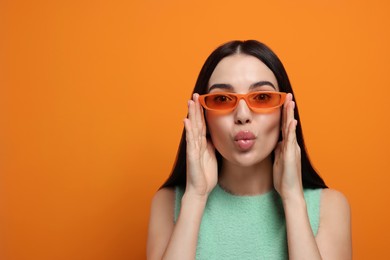 Photo of Beautiful young woman in stylish sunglasses blowing kiss on orange background. Space for text
