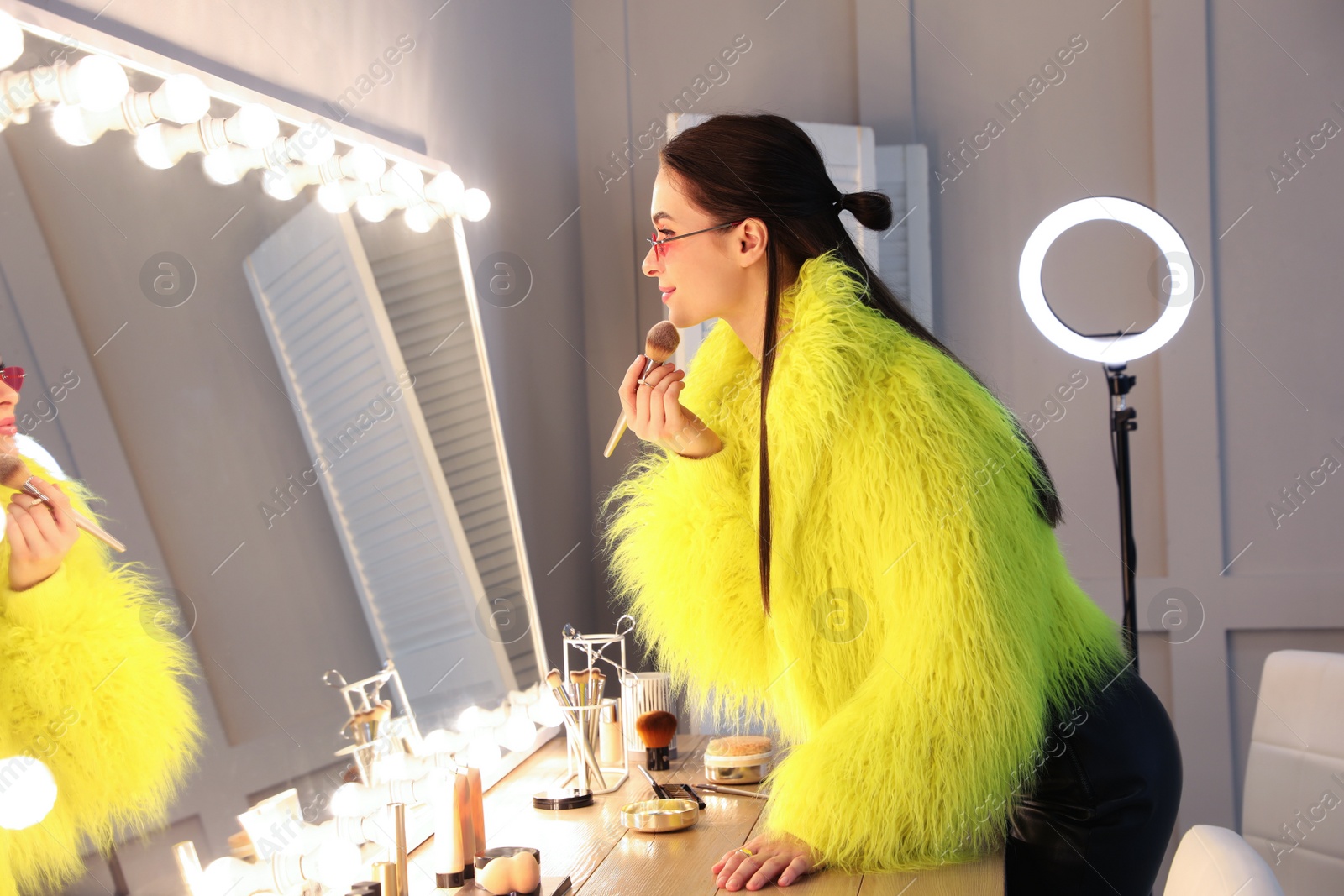 Photo of Young woman applying make up near illuminated mirror indoors