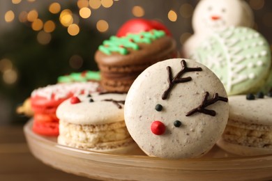 Beautifully decorated Christmas macarons on dish against blurred festive lights, closeup