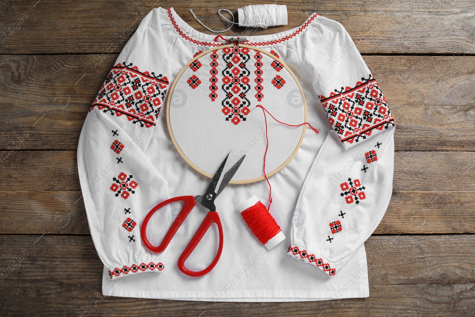 Photo of Shirt with red embroidery design in hoop, needle, scissors and threads on wooden table, flat lay. National Ukrainian clothes