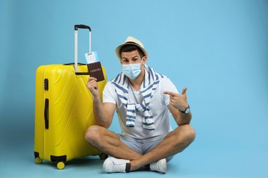 Photo of Male tourist in protective mask holding passport with ticket near suitcase on turquoise background