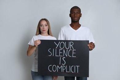 Photo of Young woman and African American man holding sign with phrase Your Silence Is Complicit on grey background. Racism concept