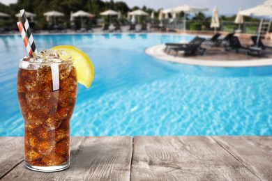 Image of Tasty refreshing soda drink with ice cubes on wooden table near outdoor swimming pool, space for text