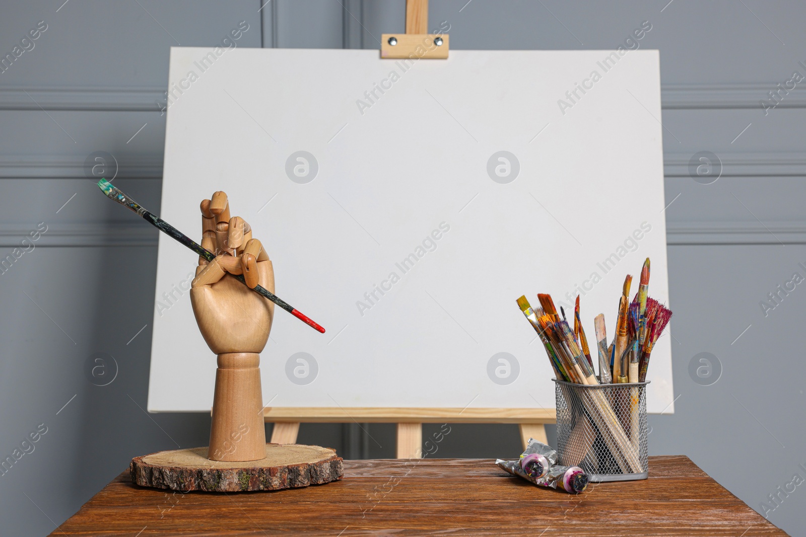 Photo of Easel with blank canvas, hand model and different art supplies on wooden table near grey wall