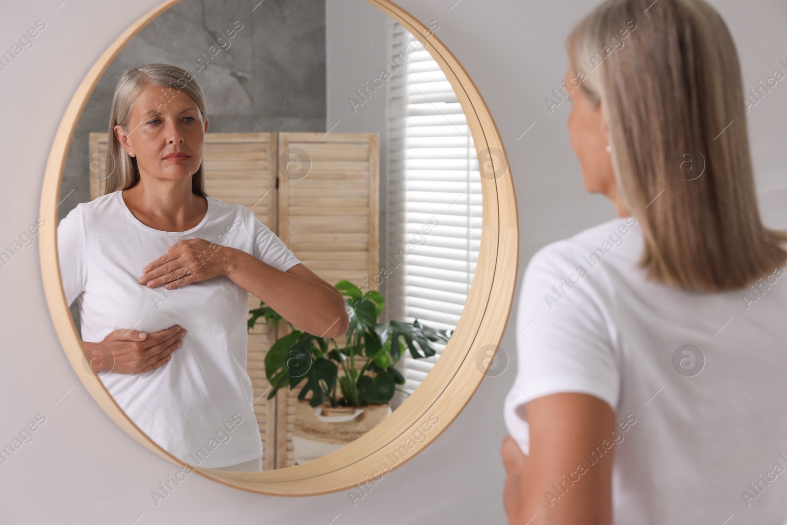 Photo of Beautiful senior woman doing breast self-examination near mirror in bathroom