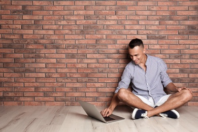 Young man sitting on floor with laptop against brick wall. Space for text