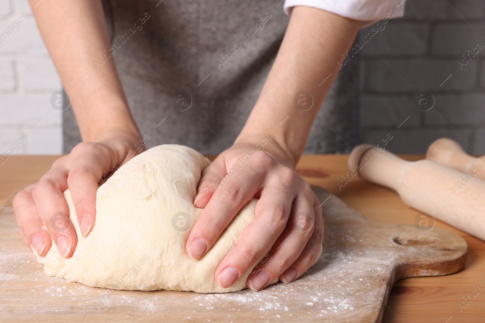 Photo of Woman kneading dough at wooden table near white brick wall, closeup