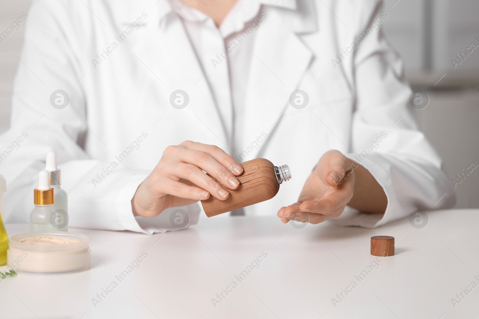 Photo of Dermatologist with bottle testing cosmetic product at white table indoors, selective focus