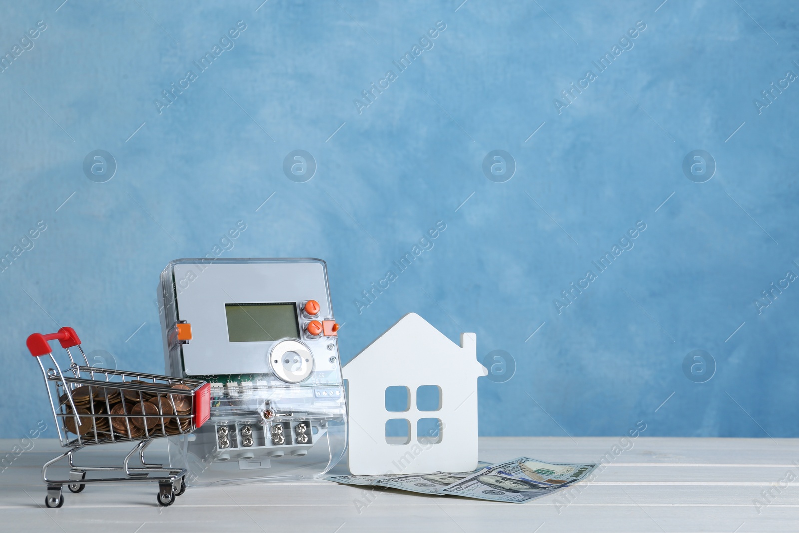 Photo of Electricity meter, house model, small shopping cart with coins and dollar banknotes on white wooden table. Space for text