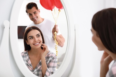 Photo of Young man presenting heart shaped balloons to his girlfriend near mirror at home