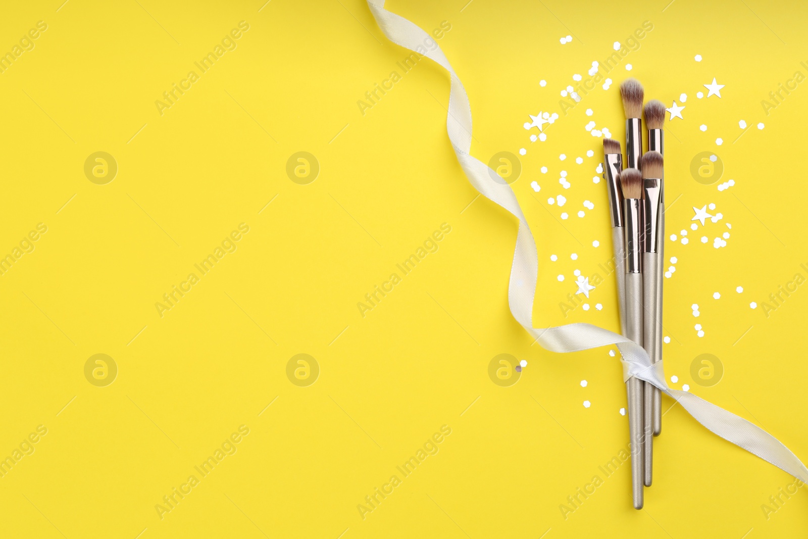 Photo of Different makeup brushes, ribbon and shiny confetti on yellow background, flat lay. Space for text