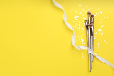 Photo of Different makeup brushes, ribbon and shiny confetti on yellow background, flat lay. Space for text