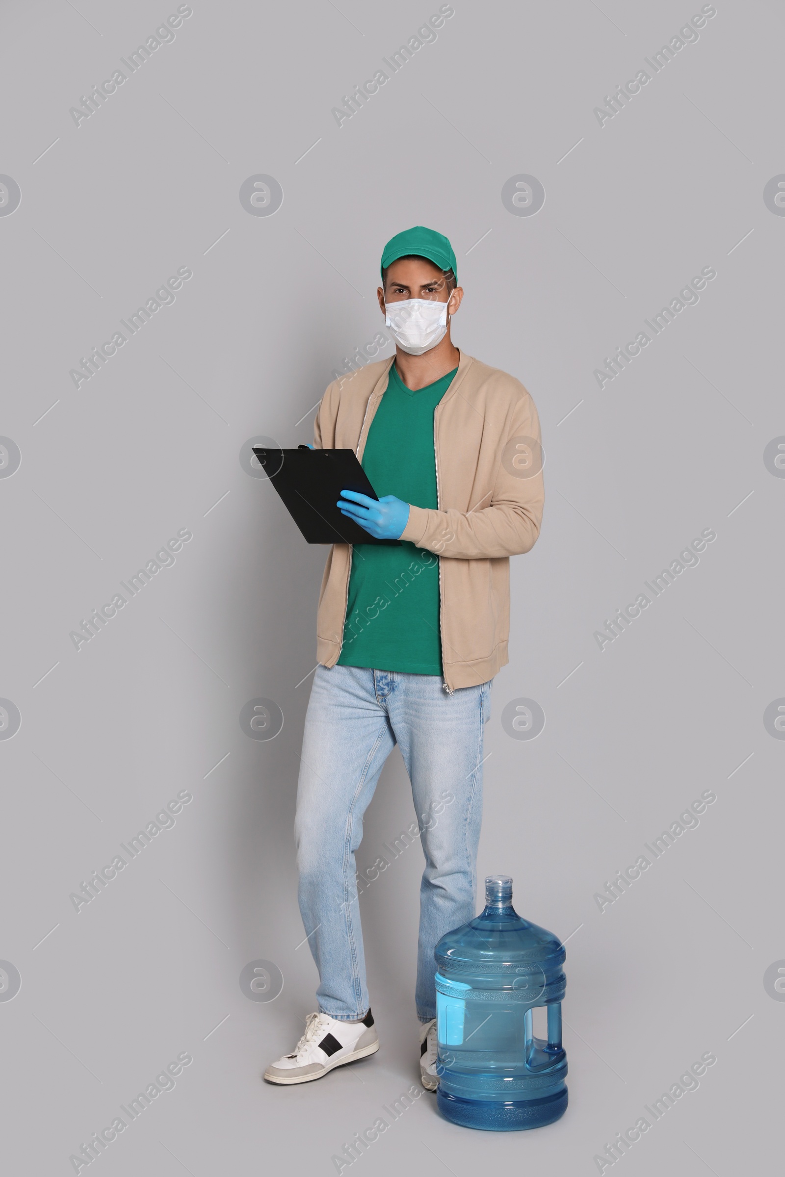 Photo of Courier in medical mask with bottle for water cooler and clipboard on light grey background. Delivery during coronavirus quarantine