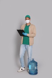 Courier in medical mask with bottle for water cooler and clipboard on light grey background. Delivery during coronavirus quarantine