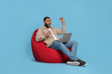 Photo of Happy young man with laptop sitting on beanbag chair against light blue background