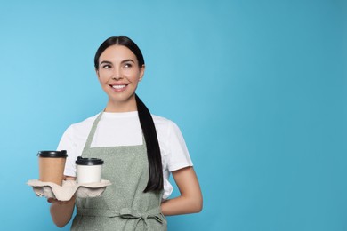 Photo of Young woman in grey apron with hot drinks on light blue background, space for text