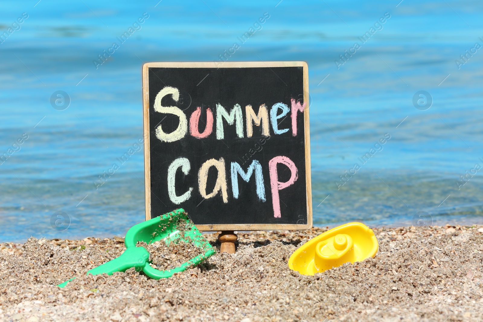 Photo of Little blackboard with text SUMMER CAMP and toys on sand near sea