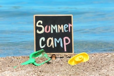 Photo of Little blackboard with text SUMMER CAMP and toys on sand near sea