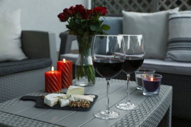 Photo of Glasses of wine, vase with roses, burning candles and snacks on rattan table at balcony in evening, closeup