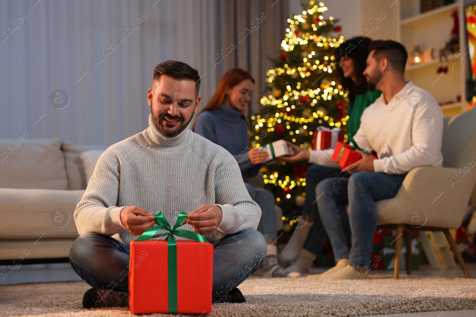 Photo of Christmas celebration in circle of friends. Happy man opening gift at home, selective focus