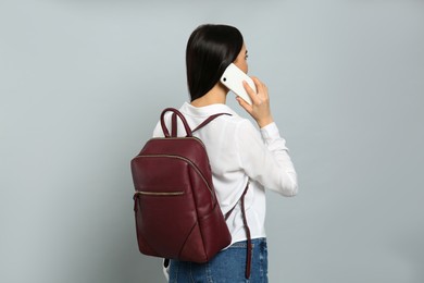 Young woman with stylish backpack talking on phone against light grey background, back view