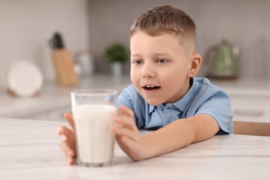 Photo of Cute boy reaching out for glass of milk at white table in kitchen