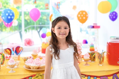 Cute little girl near table with treats at birthday party indoors