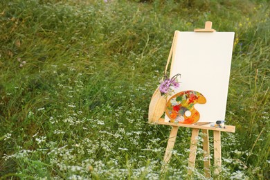 Wooden easel with blank canvas, painting equipment and flowers in meadow. Space for text