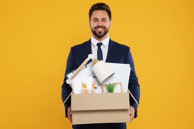 Happy unemployed man with box of personal office belongings on orange background