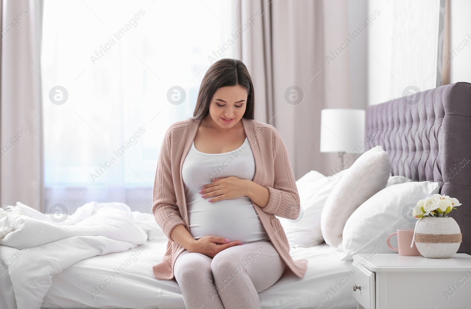 Photo of Young beautiful pregnant woman sitting on bed and touching her belly at home