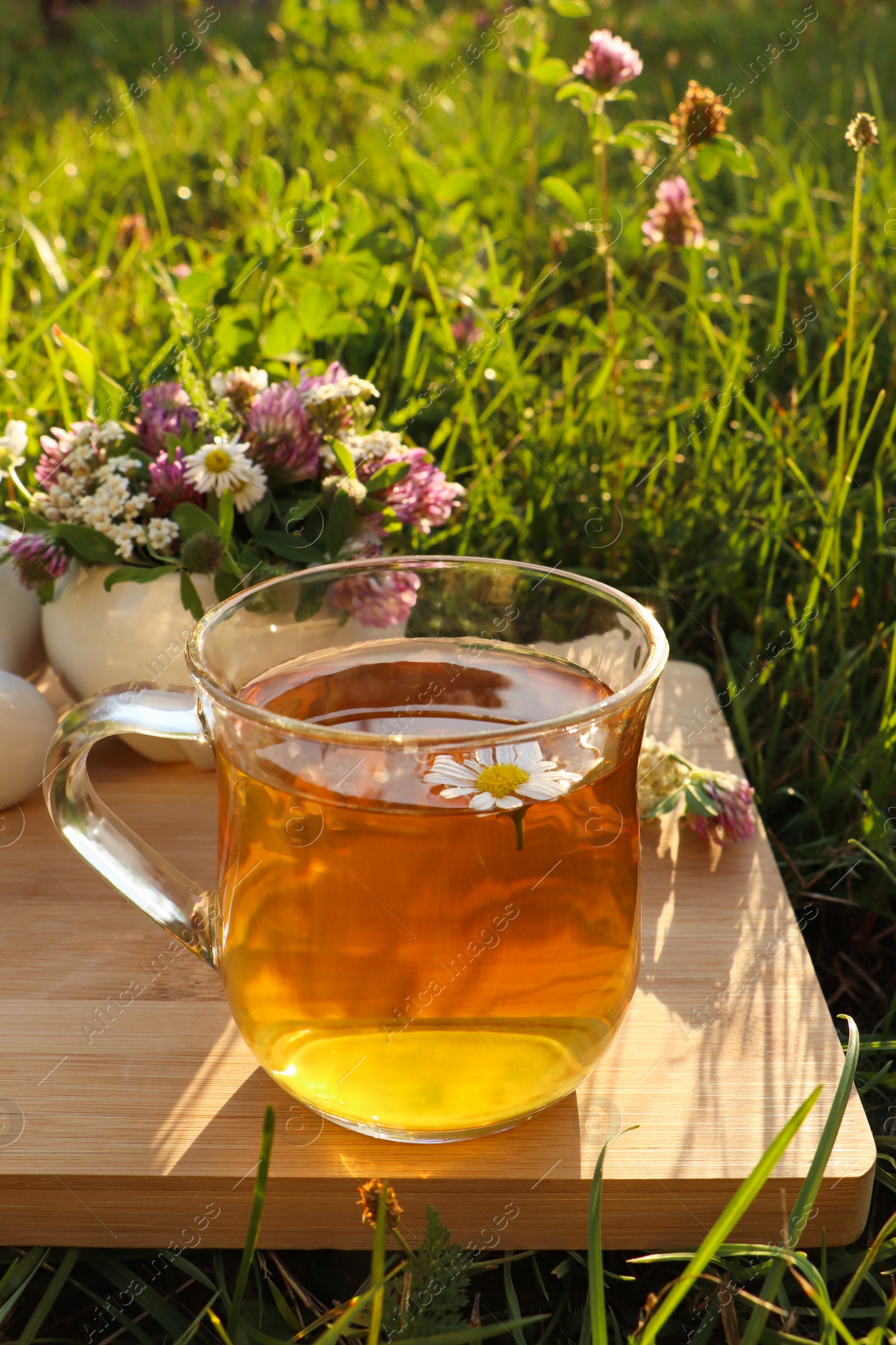 Photo of Cup of aromatic herbal tea, pestle and ceramic mortar with different wildflowers on wooden board in meadow