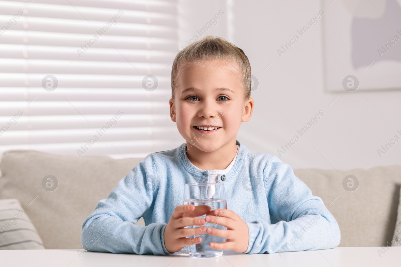 Photo of Happy little girl holding glass of fresh water at white table indoors