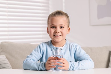 Happy little girl holding glass of fresh water at white table indoors