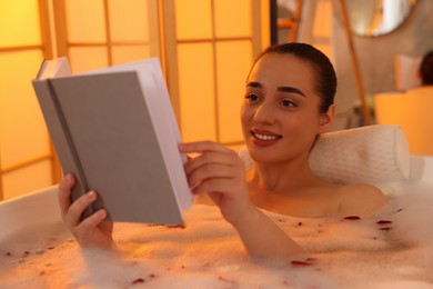 Photo of Happy woman reading book while taking bath in tub with foam and rose petals indoors