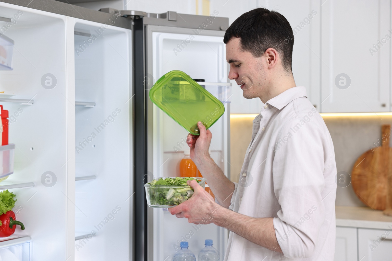 Photo of Happy man holding container with vegetables near refrigerator in kitchen