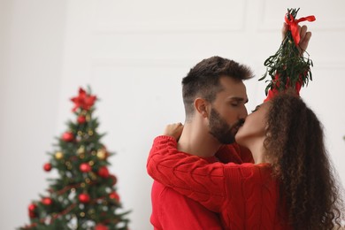 Photo of Happy couple kissing under mistletoe bunch in room decorated for Christmas