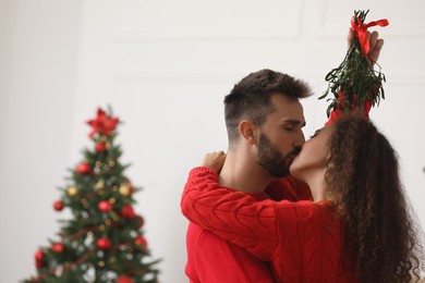 Happy couple kissing under mistletoe bunch in room decorated for Christmas