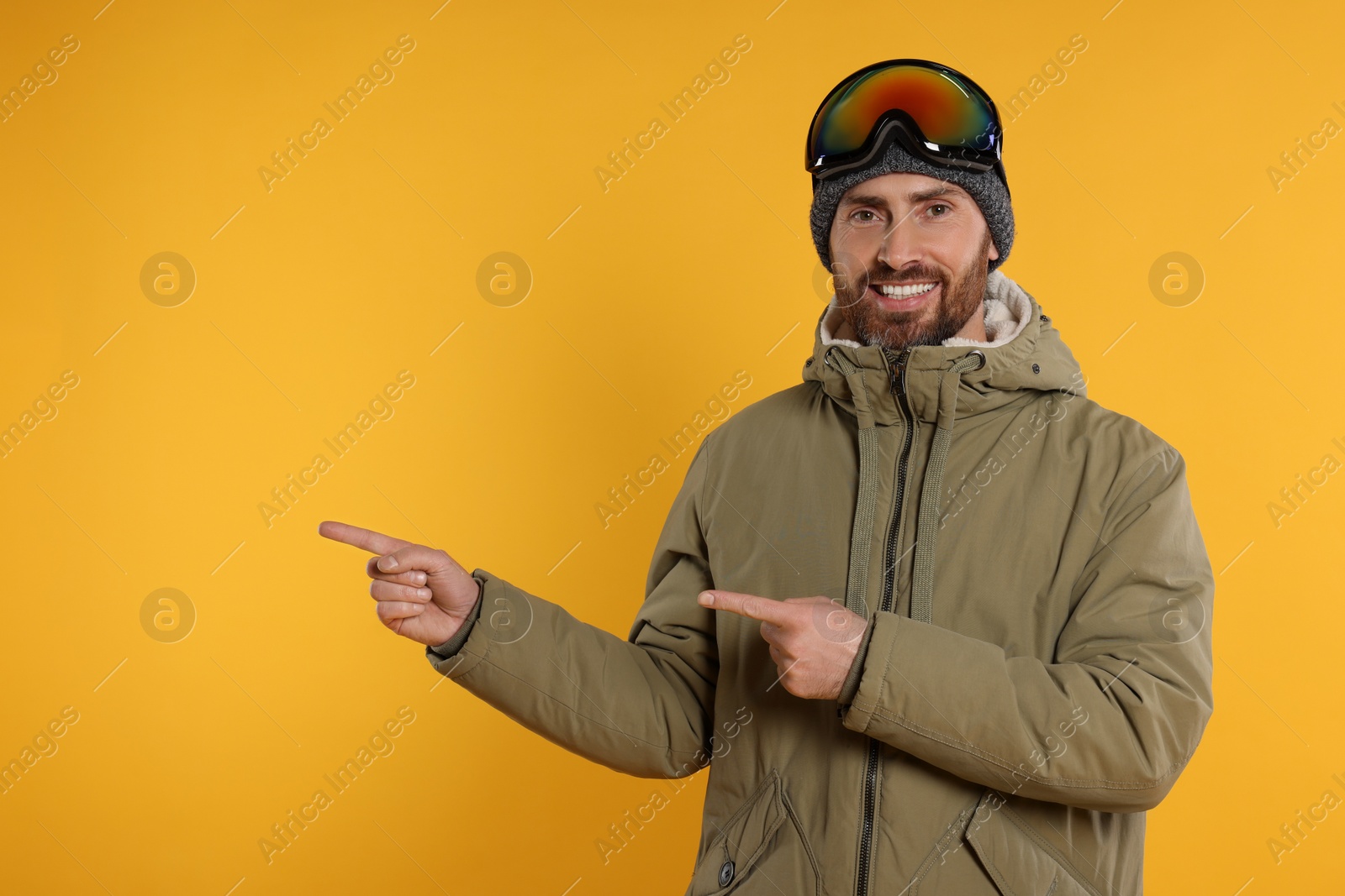 Photo of Winter sports. Happy man in ski suit and goggles pointing at something on orange background, space for text