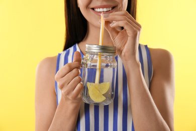 Photo of Young woman drinking tasty lemon water on yellow background, closeup