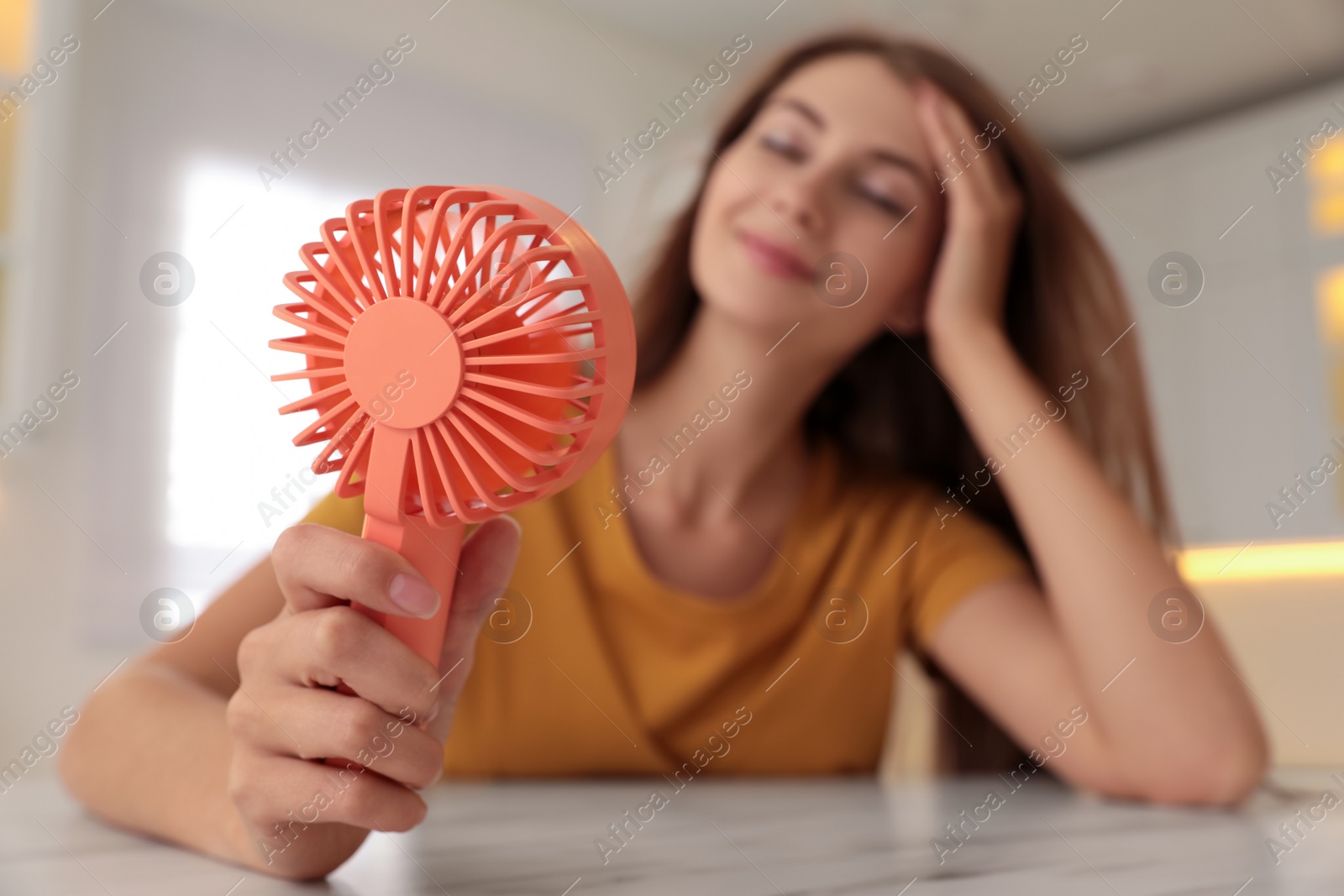 Photo of Woman enjoying air flow from portable fan at table in kitchen, focus on device. Summer heat