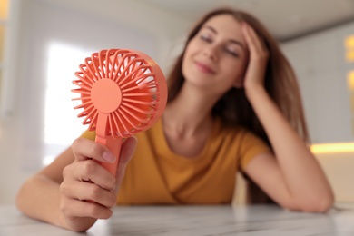 Woman enjoying air flow from portable fan at table in kitchen, focus on device. Summer heat