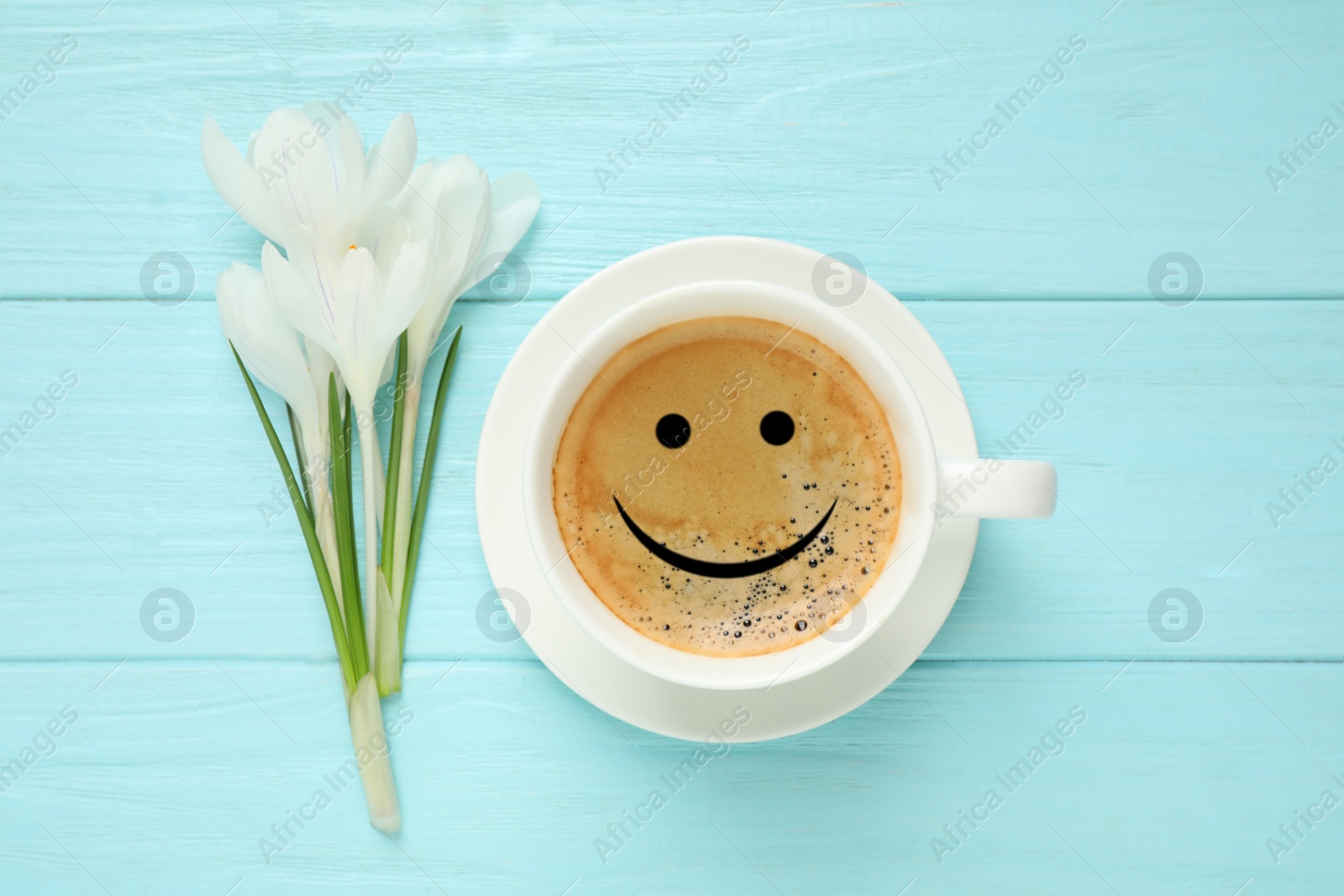 Image of Cup of aromatic coffee with happy face and crocuses on light blue wooden table, flat lay. Good morning