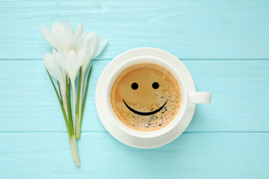 Cup of aromatic coffee with happy face and crocuses on light blue wooden table, flat lay. Good morning