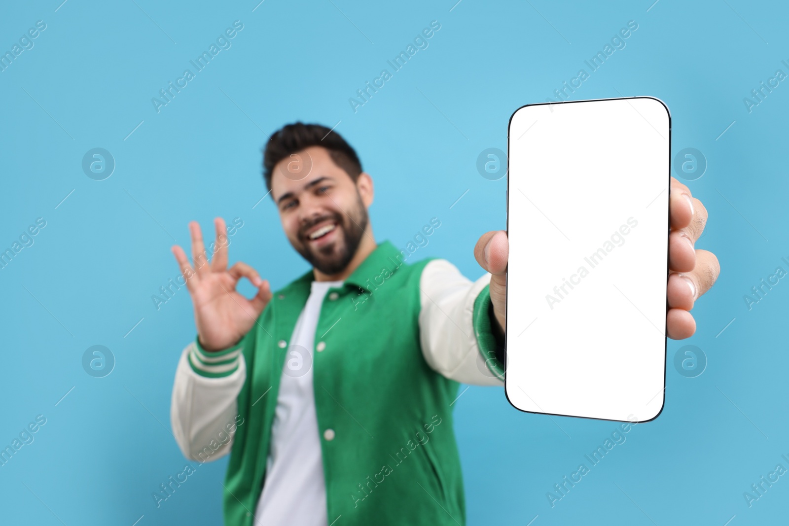 Photo of Young man showing smartphone in hand and OK gesture on light blue background, selective focus. Mockup for design