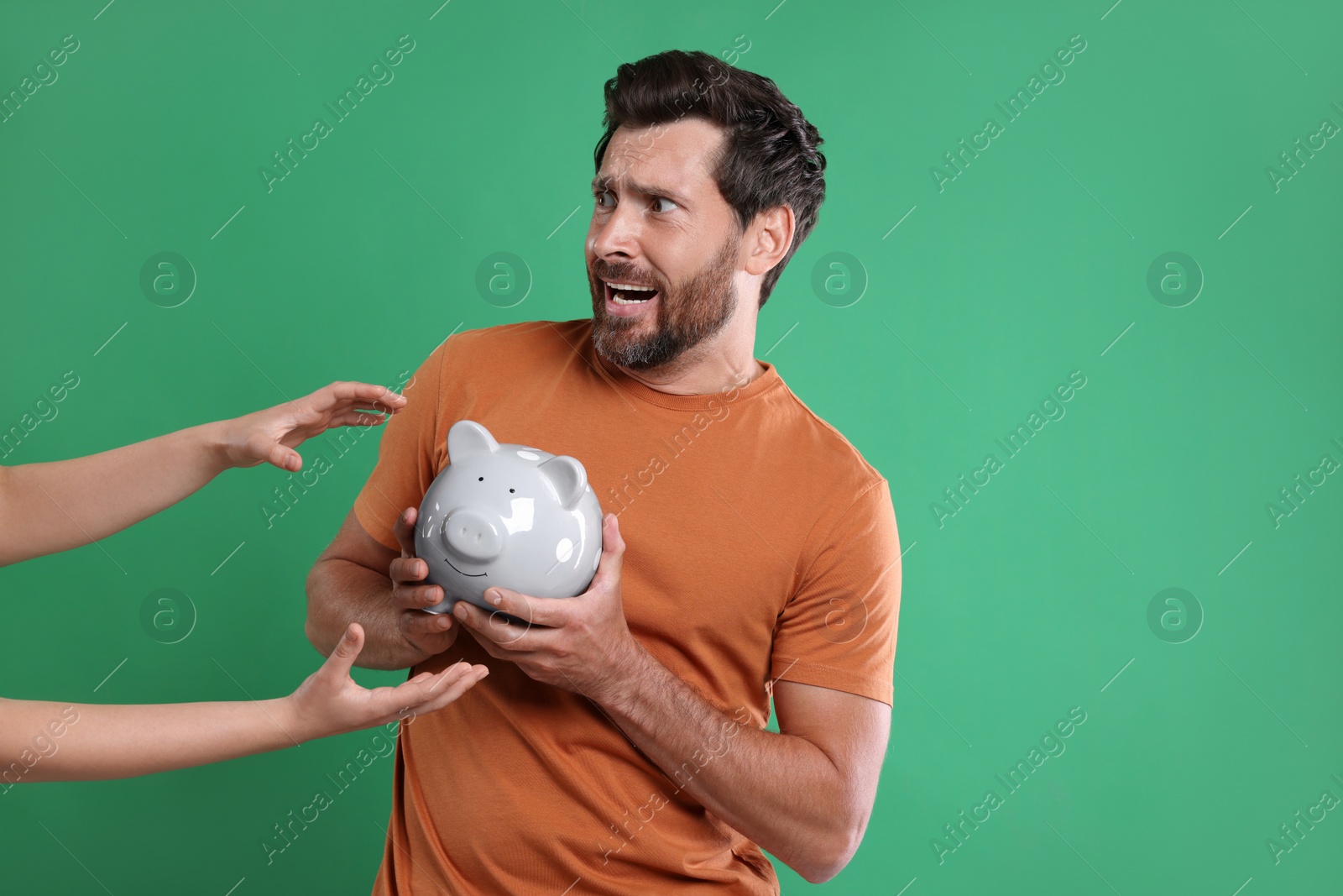 Photo of Scared man trying to protect piggy bank from woman on green background, space for text. Be careful - fraud