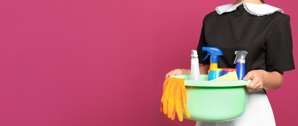 Image of Young chambermaid holding plastic basin with detergents on color background, closeup view with space for text. Banner design