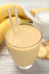 Glass of tasty banana smoothie with straws and ingredients on white wooden table, closeup