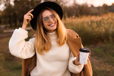 Photo of Beautiful young woman with cup of coffee wearing stylish autumn clothes outdoors