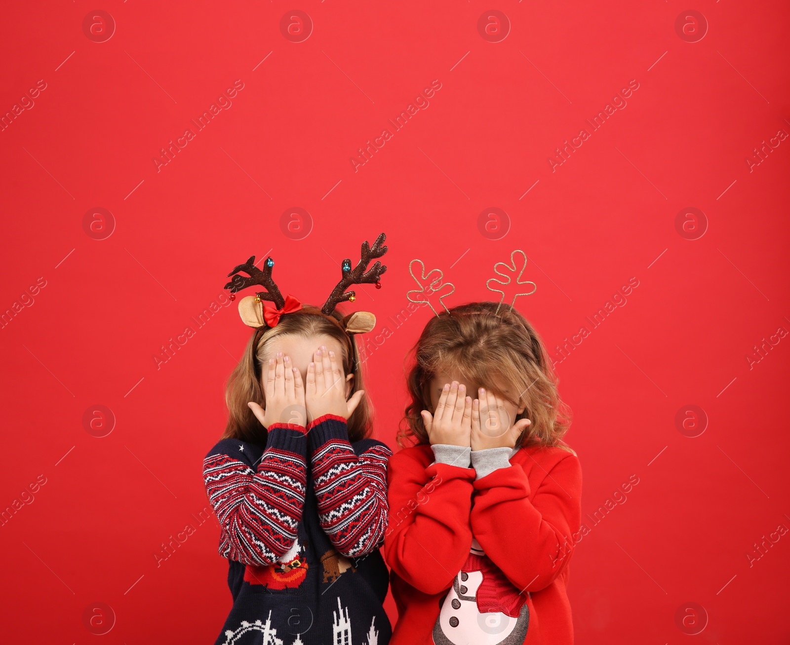 Photo of Kids in Christmas sweaters and festive headbands hiding faces on red background